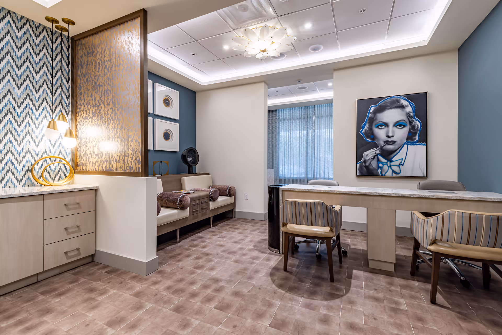 A modern interior room with a long table and four chairs, two on each side. On the left side, there is a built-in cabinet with drawers and a decorative wall with hanging lights and a golden sculpture. The back wall features a bench with cylindrical pillows and framed artwork above it. On the right wall, there is a large portrait of a woman with blue makeup. The room has a tiled floor and a ceiling with recessed lighting and a decorative light fixture.