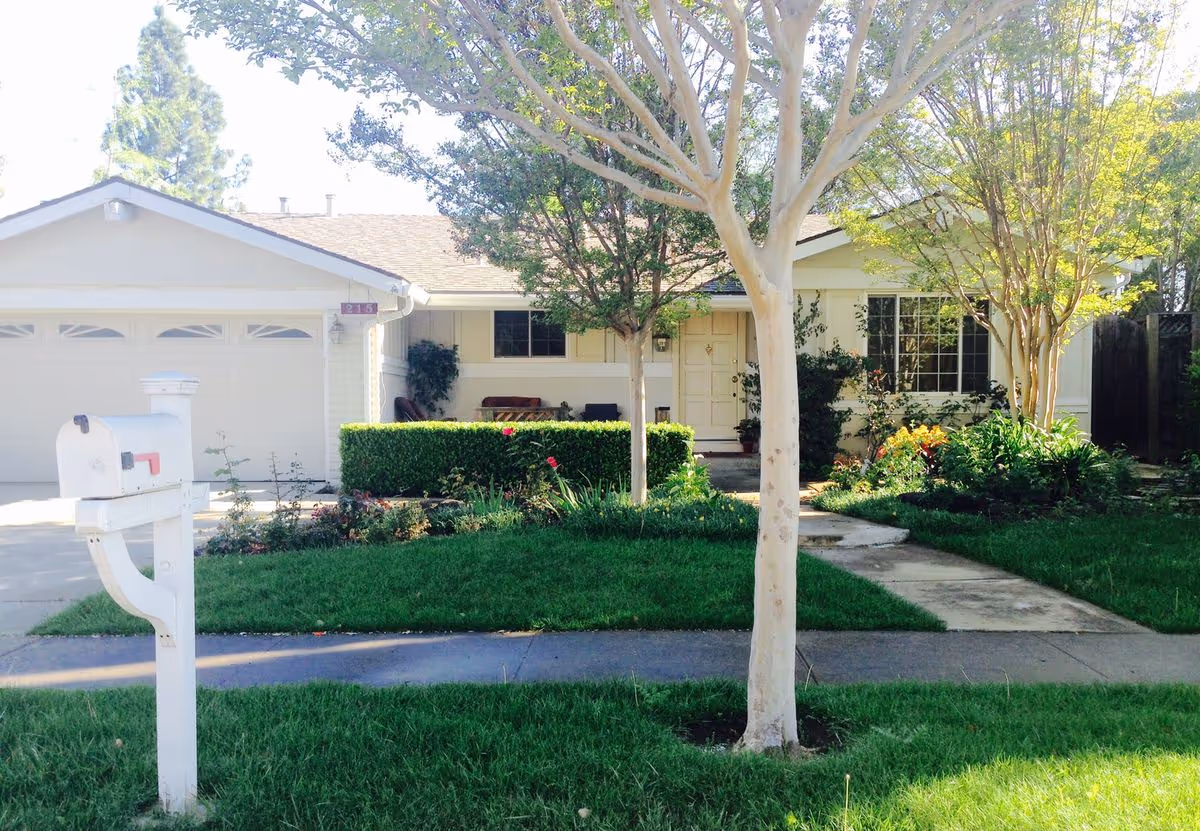 Front of a single-story suburban house with a garage, mailbox, walkway, trees, and a manicured front yard.