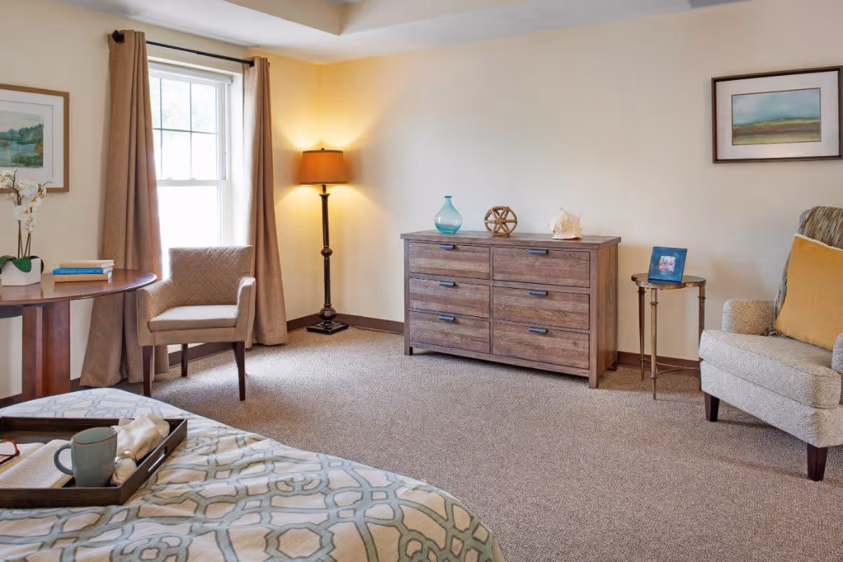 A cozy bedroom with a patterned bedspread, a wooden dresser topped with decorative items, a cushioned armchair with a yellow pillow, a small round side table with a framed photo, a floor lamp, a window with beige curtains, and a round wooden table with a chair and books.
