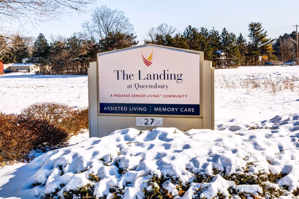 Outdoor view of a sign for The Landing at Queensbury, a Pegasus Senior Living community offering assisted living and memory care services. The sign is surrounded by snow-covered bushes and a snowy field with trees in the background.