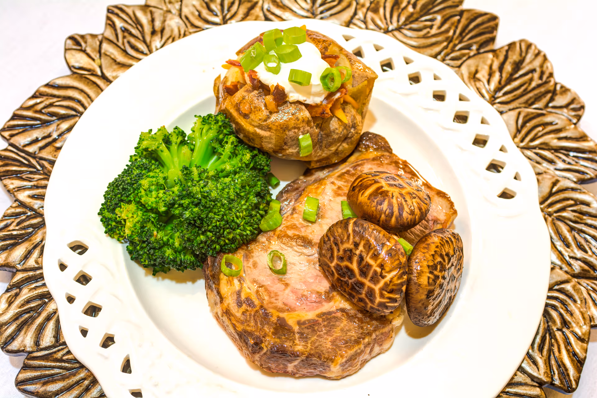 A plated meal consisting of a baked potato topped with sour cream and chopped green onions, steamed broccoli, and grilled mushrooms served on a white decorative plate with a bronze-colored ornate charger underneath.