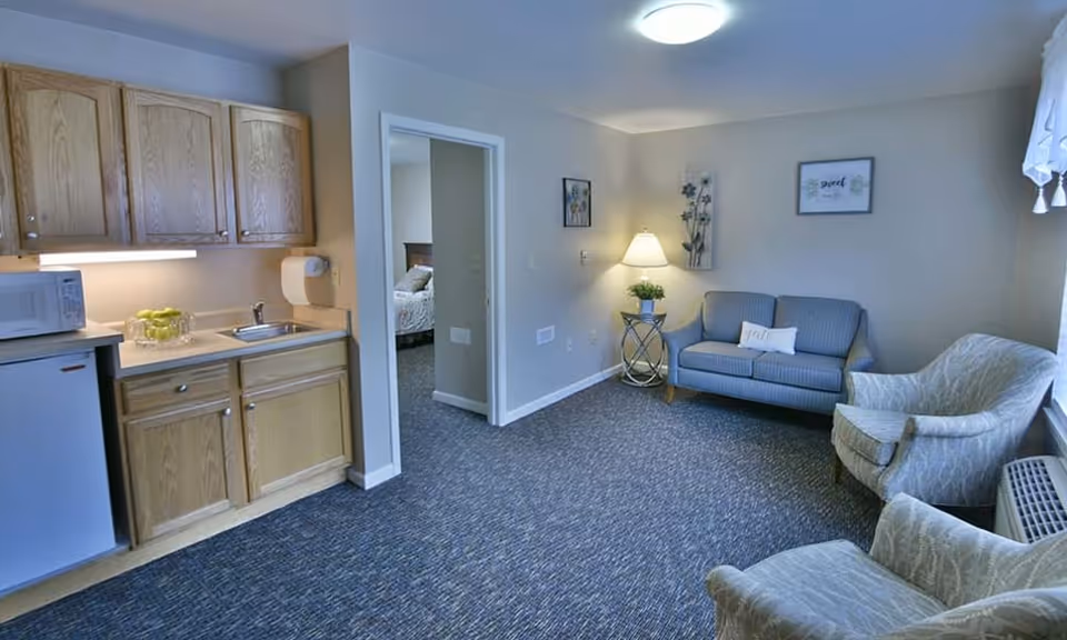 A cozy assisted living apartment interior featuring a small kitchenette with wooden cabinets, a microwave, mini fridge, and a sink on the left. To the right is a sitting area with a blue loveseat, two patterned armchairs, a side table with a lamp and plant, and framed artwork on the walls. A doorway leads to a bedroom with a bed visible in the background.