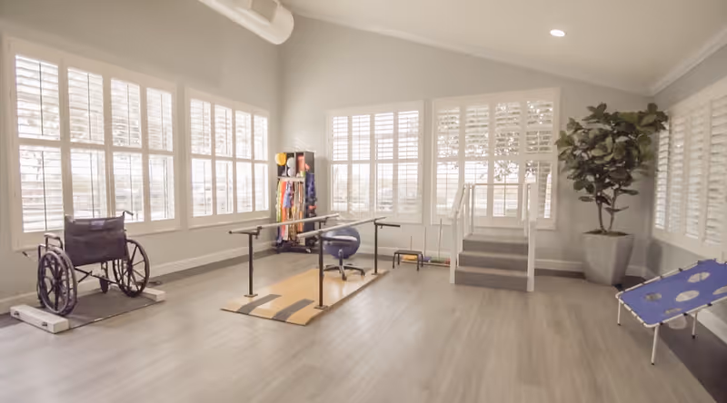 Bright rehabilitation room with parallel bars, a wheelchair, exercise equipment, and large shuttered windows.