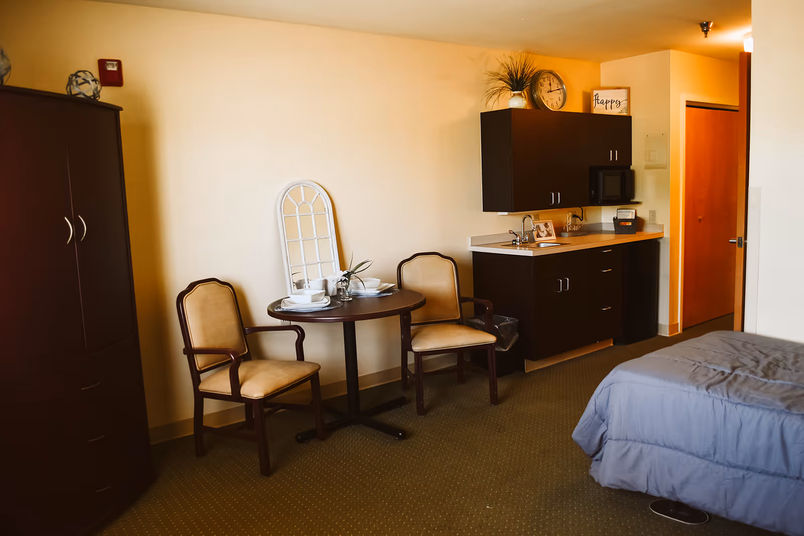 Studio bedroom with a bed, a round table and two chairs, and a small kitchenette with dark cabinets.