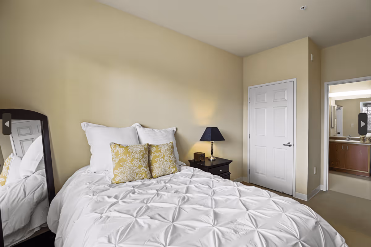 A bedroom with a neatly made bed featuring white bedding and two yellow patterned pillows. There is a dark wooden nightstand with a lamp beside the bed. A full-length mirror is positioned on the left side of the bed, reflecting part of the room. A closed white door is visible on the right wall, and an open doorway leads to a bathroom with a sink and mirror.