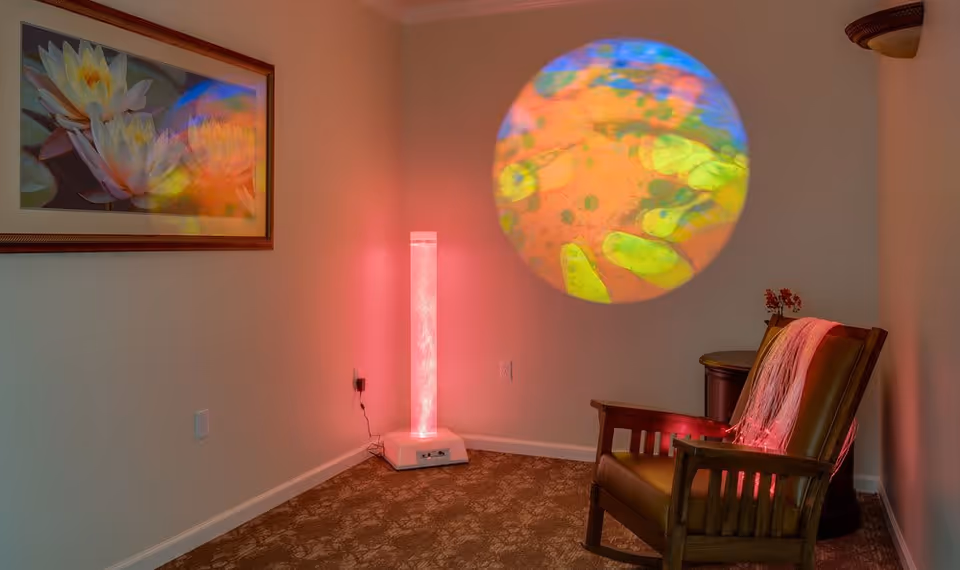 A cozy corner of a room with a wooden armchair draped with a pink scarf, a small round table with a flower vase, and a tall cylindrical light emitting a soft pink glow. A colorful circular projection is displayed on the wall above the chair, and a framed picture of white flowers hangs on the adjacent wall.