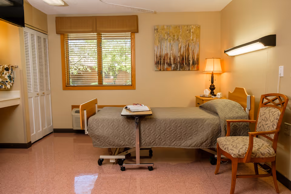 A single bed with a gray quilted bedspread in a senior living facility room. There is a wooden chair with patterned upholstery next to the bed, a small bedside table with a lamp, tissues, and a phone. A window with wooden blinds and a valance is on the wall behind the bed, and a painting of trees hangs above the bedside table. The floor is a polished pinkish tile, and there is a white closet with louvered doors on the left side of the room.
