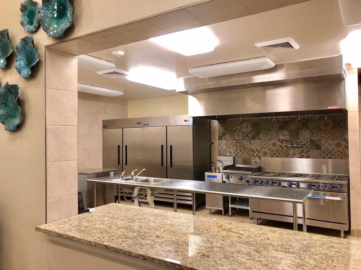A commercial kitchen with stainless steel appliances including a large stove with multiple burners, a stainless steel sink, and large refrigerators. The backsplash features decorative patterned tiles, and there is a granite countertop in the foreground. The kitchen is well-lit with ceiling lights and has beige tiled walls.