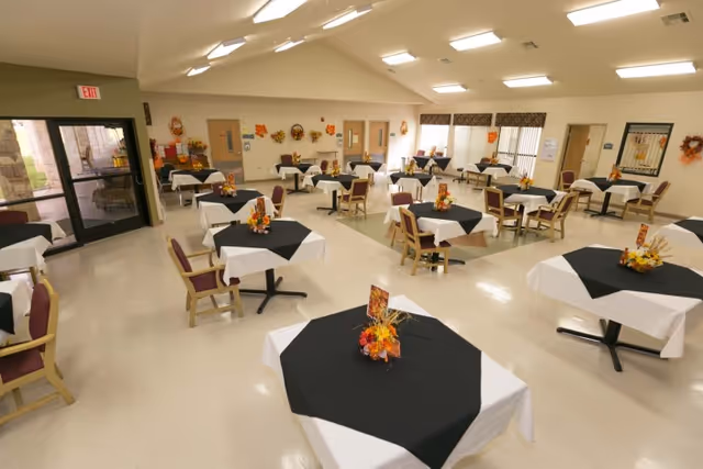 A spacious dining room with multiple square tables covered with white tablecloths and black overlays, each decorated with a small autumn-themed floral centerpiece. The room has beige walls adorned with fall wreaths and decorations, several wooden chairs with purple cushions around the tables, and large windows letting in natural light. There is a glass door exit on the left side of the room.