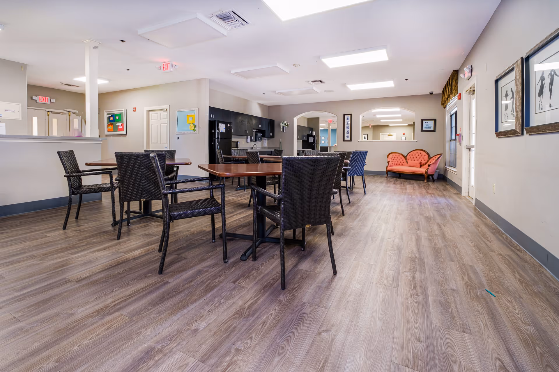 A spacious common area with wooden flooring featuring several dark wicker chairs and tables. In the background, there is a kitchenette with black cabinets and appliances. On the far side, a coral-colored loveseat is placed against the wall. The walls are decorated with framed artwork, and there are multiple ceiling lights providing bright illumination.