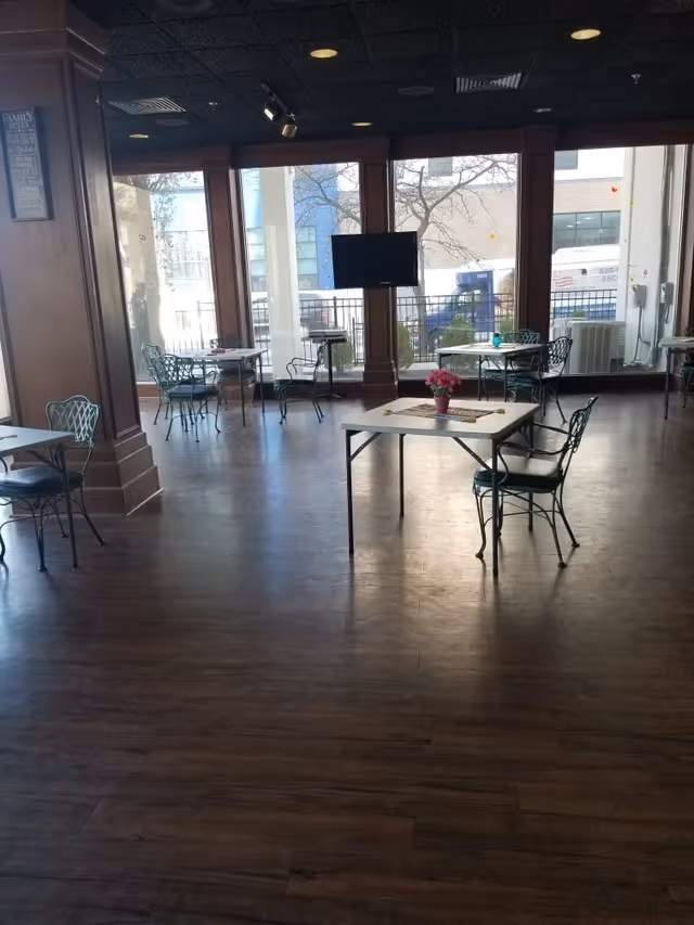 A spacious room with wooden flooring and several metal tables and chairs arranged throughout. Each table has a small centerpiece, such as a flower pot. Large windows line the back wall, allowing natural light to fill the room and providing a view of the outside. A flat-screen TV is mounted on a wooden pillar near the windows.