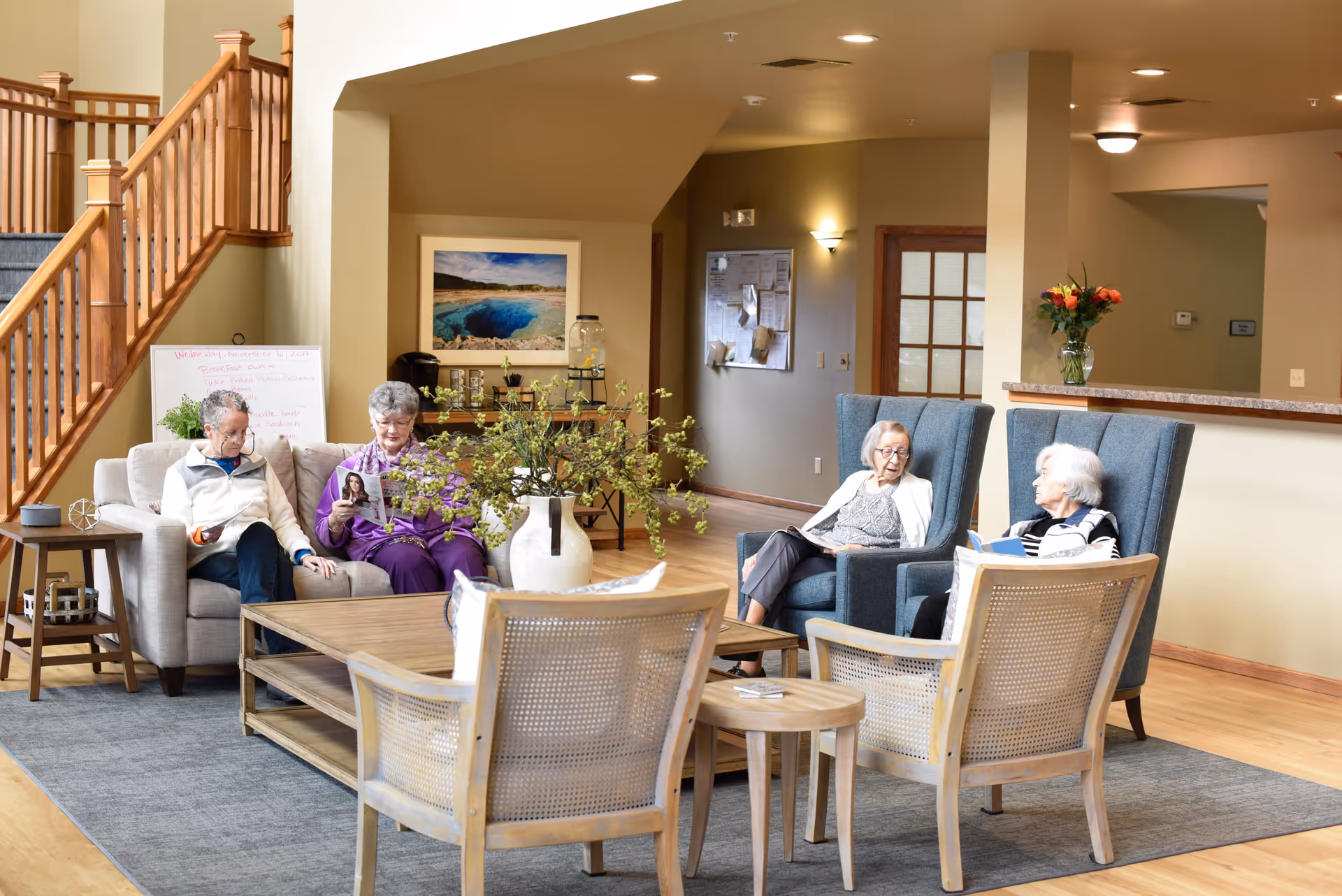 Four senior residents sitting and chatting in a well-lit assisted-living common lounge with armchairs, a coffee table, rug, and wooden staircase.