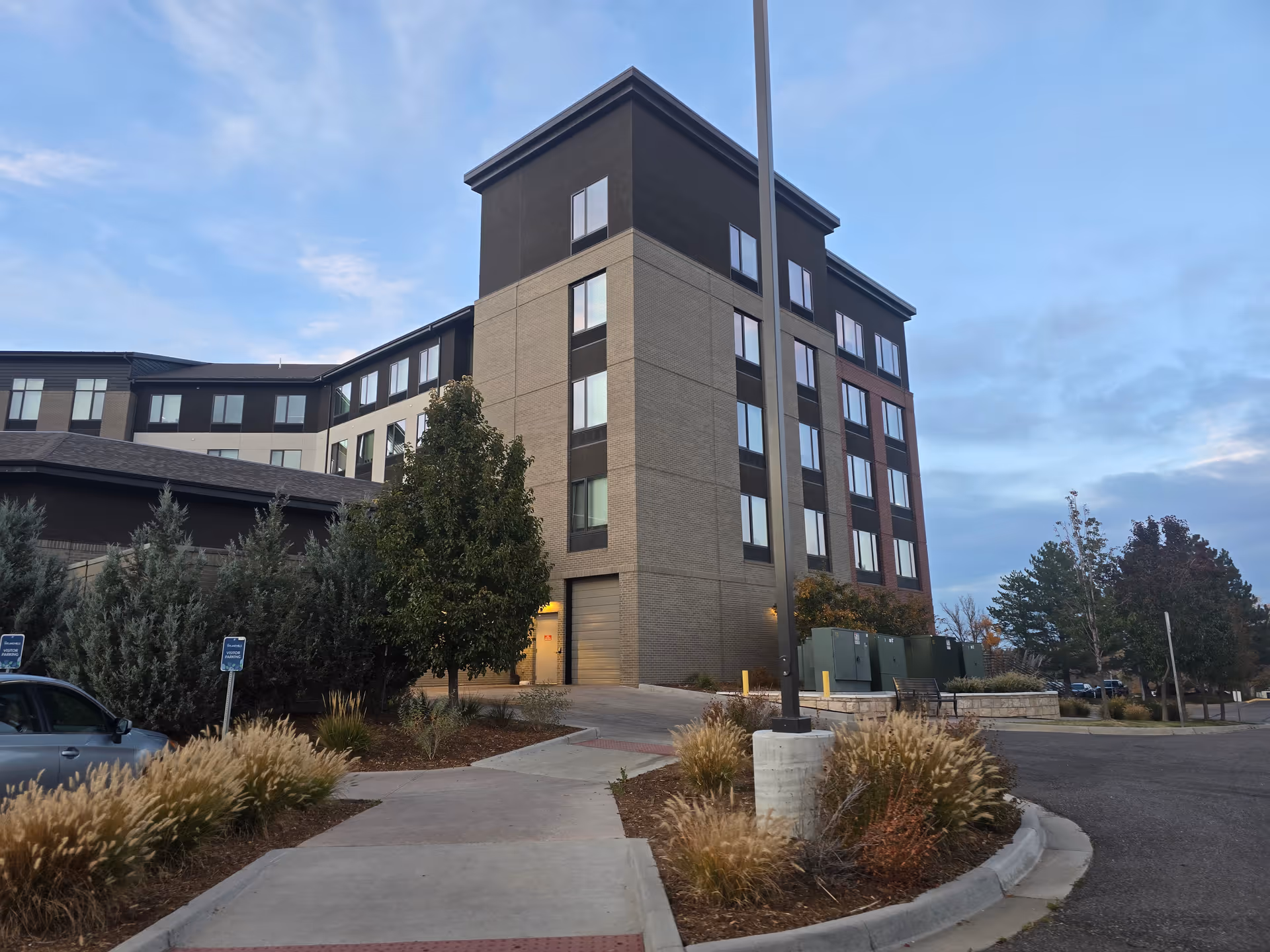 Exterior view of a multi-story senior living facility building with beige and dark brown brick facade, surrounded by landscaped bushes, trees, and a parking area under a partly cloudy sky.