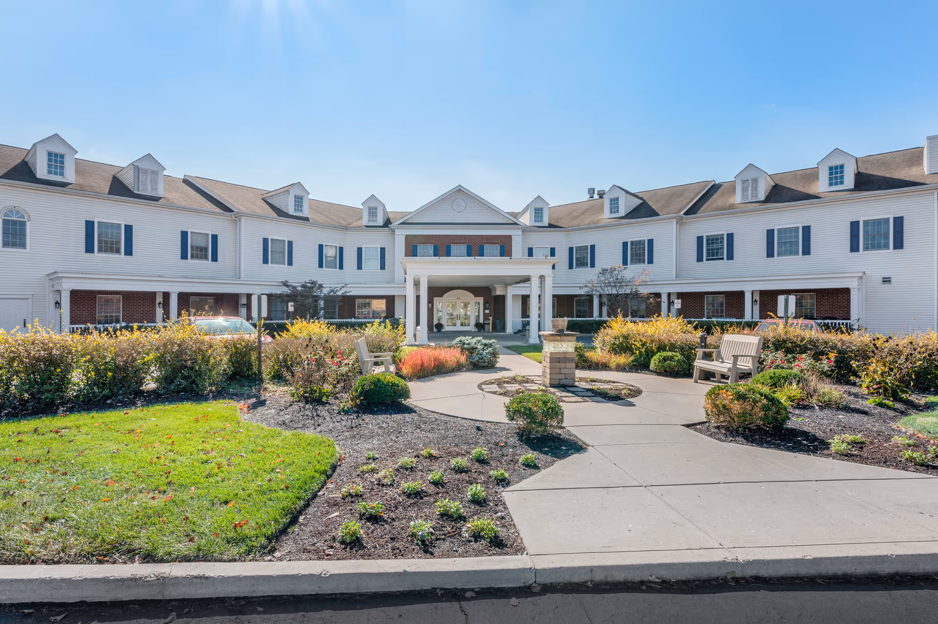 Exterior view of a large two-story senior living facility building with white siding and multiple dormer windows. The entrance features a covered porch with columns, and there is a landscaped garden area with bushes, flowers, and benches in front of the building under a clear blue sky.