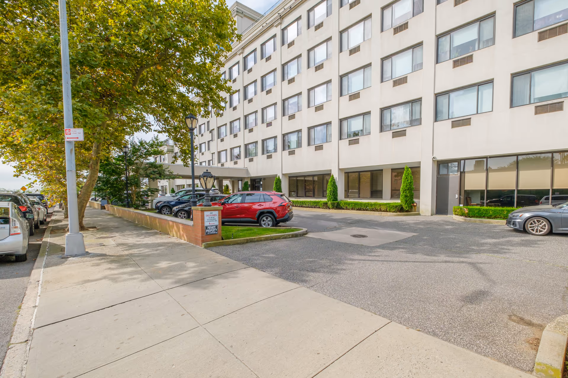 Exterior view of a multi-story senior living facility building with several windows, a driveway with parked cars, a sidewalk with trees, and a small landscaped area with bushes and a sign near the entrance.