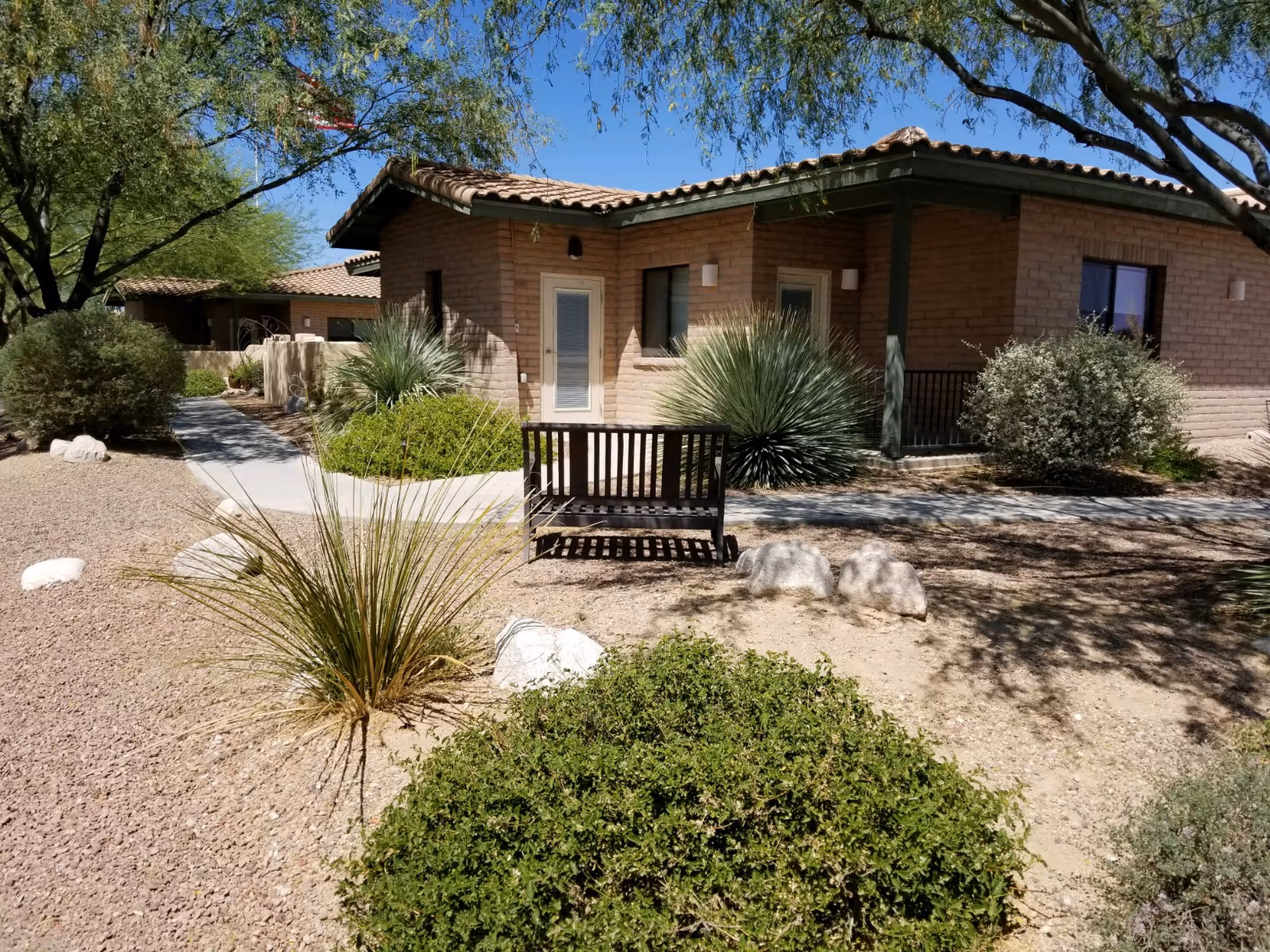 Outdoor view of a single-story brick building with a tiled roof at BridgeWater Assisted Living - Tucson. The scene includes desert landscaping with bushes, rocks, and trees, a concrete pathway, and a wooden bench in the foreground under clear blue sky.