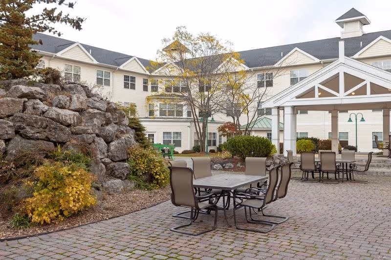 Outdoor patio area at Legacy at Clover Blossom with several tables and chairs on a brick-paved surface, surrounded by landscaping including rocks, bushes, and trees, with a multi-story building in the background.