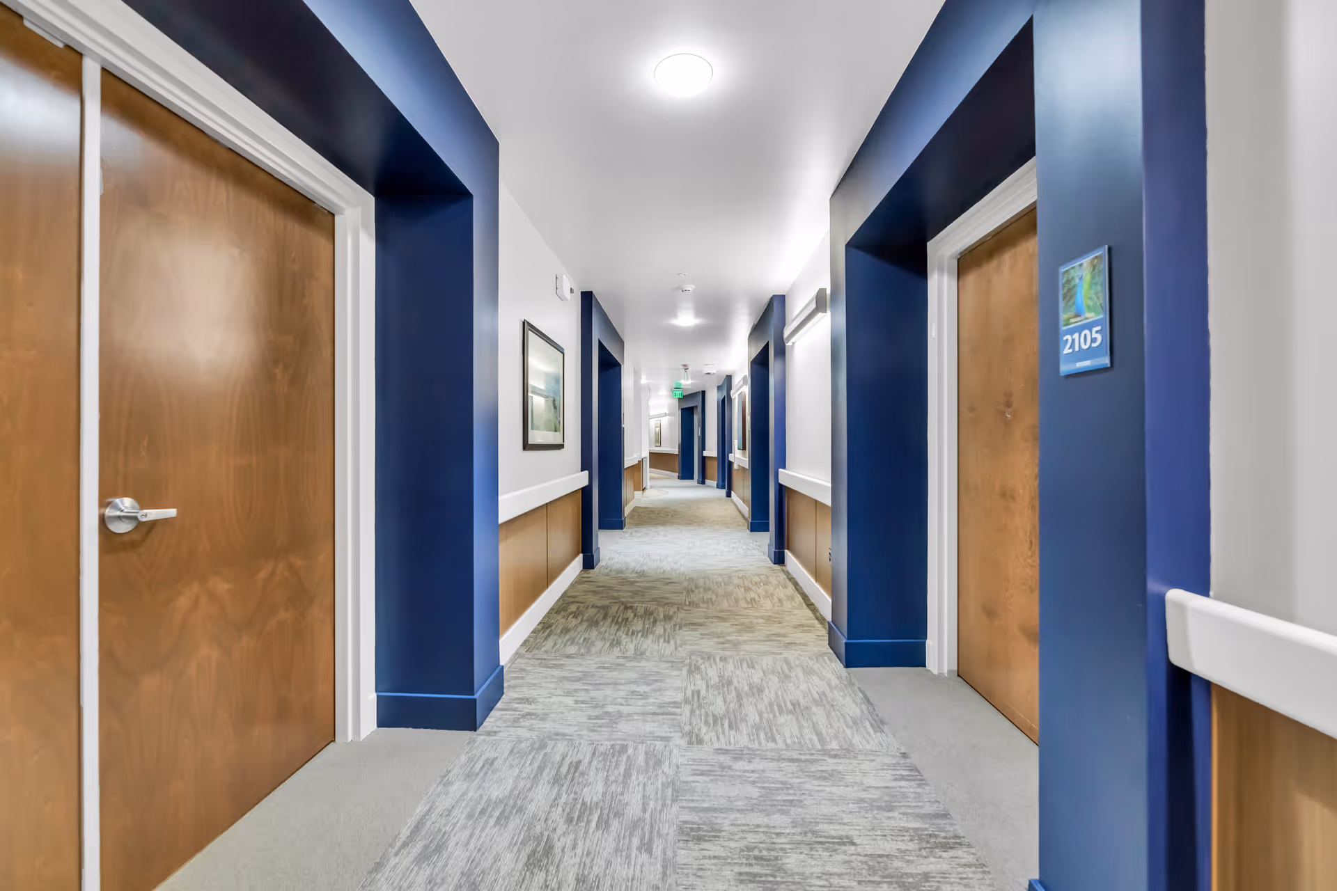 A well-lit hallway in a senior living facility with blue and white walls, wooden doors on both sides, and carpeted flooring. The hallway features framed artwork on the walls and room number signs next to the doors.