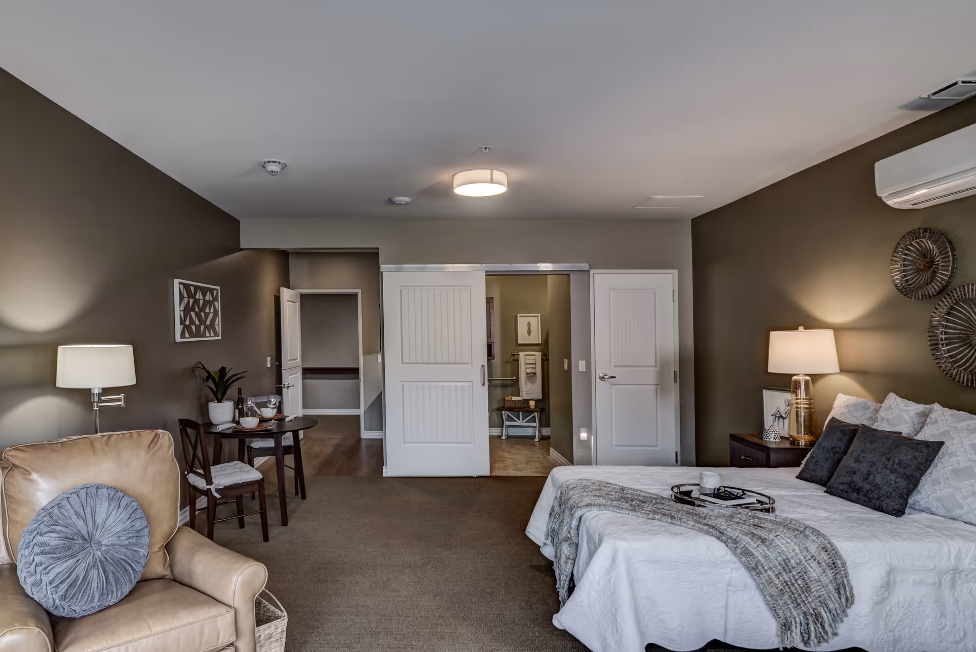 A cozy bedroom in an assisted living facility featuring a bed with white bedding and dark pillows, a beige armchair with a round gray cushion, a small dining table set for two, and a view into a bathroom with sliding white doors. The walls are painted in neutral tones, and the room is softly lit by lamps on either side of the bed.