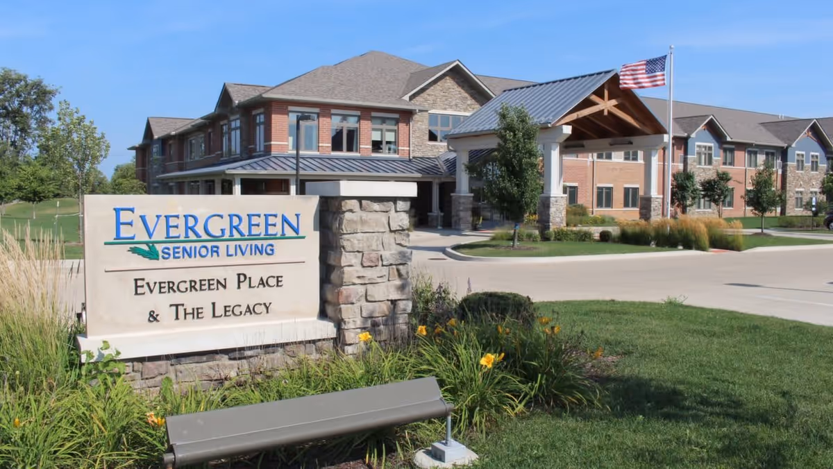Exterior view of Evergreen Senior Living facility showing a large brick and stone building with multiple windows, a covered entrance with wooden beams, an American flag on a flagpole, and a stone sign in the foreground that reads 'Evergreen Senior Living Evergreen Place & The Legacy' surrounded by green grass and flowering plants.