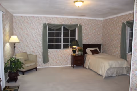 A bedroom with floral wallpaper, a single bed with beige bedding and pillows, a wooden nightstand with a lamp and a plant, a beige armchair, a floor lamp, a potted plant, and a window with green curtains.