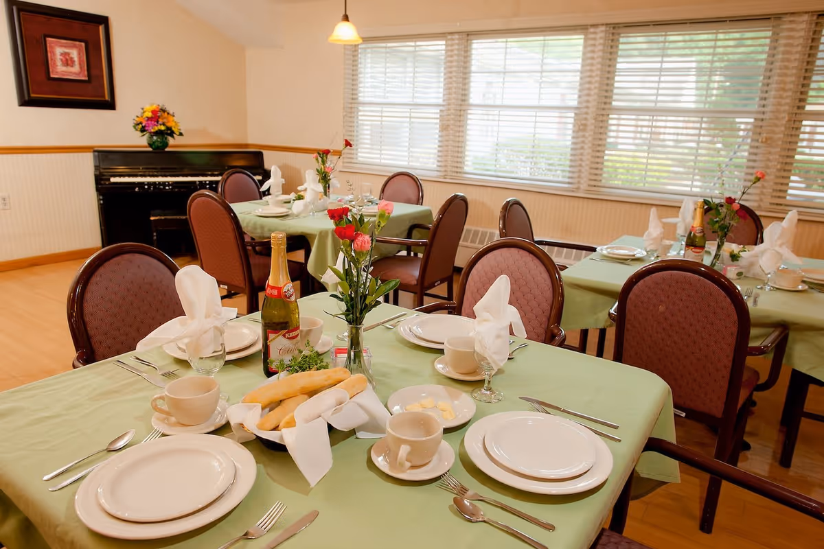 A dining room set up with tables covered in green tablecloths, each set with white plates, cups, silverware, and folded white napkins. Breadsticks in a basket, small plates with butter, and bottles of sparkling cider are on the tables. There are chairs with maroon upholstery around the tables, a piano against the wall, a framed picture, and large windows with blinds letting in natural light.