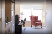 A bright room with a large window covered by white blinds. In front of the window is a red upholstered armchair and a wooden desk with a chair and a white desk lamp. The room has light-colored walls and carpeted floor.