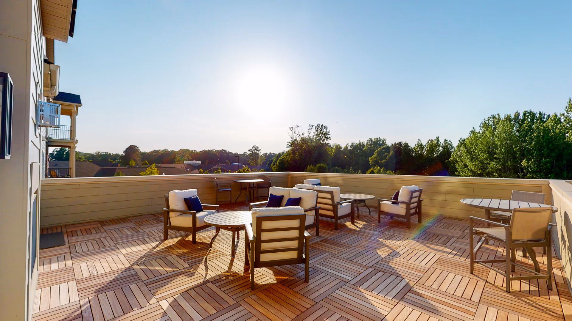 Outdoor patio area with wooden tile flooring, several cushioned chairs with dark frames, round tables, and a clear blue sky with the sun shining brightly. Trees and rooftops are visible in the background.