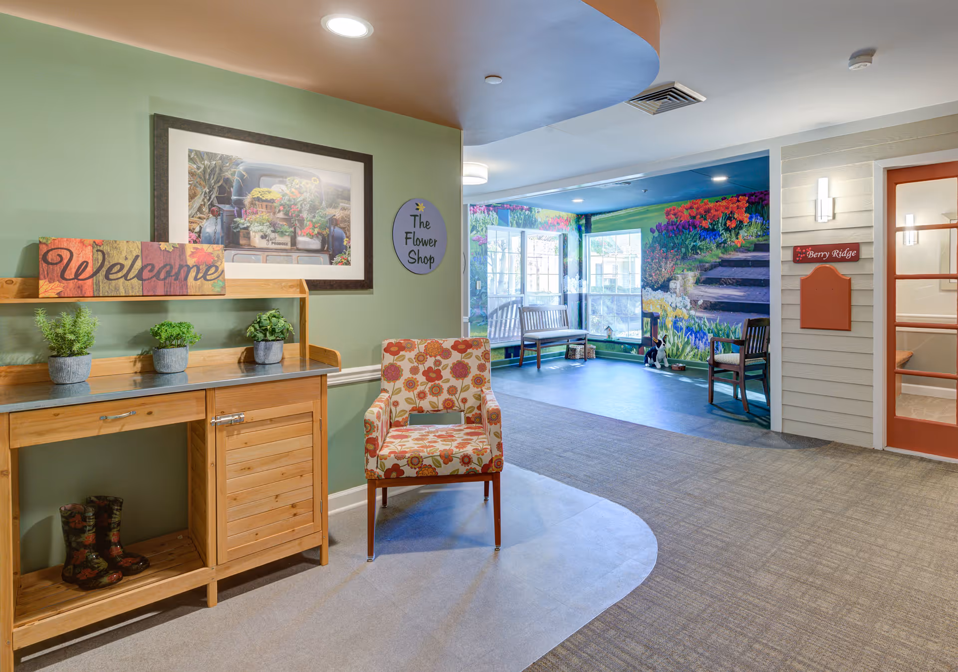 Interior view of a senior living facility area with a wooden console table holding three small potted plants and a colorful 'Welcome' sign. A floral patterned armchair is placed nearby. The wall behind has a framed picture of flowers and a round sign that reads 'The Flower Shop'. In the background, there is a seating area with benches and chairs, and a large mural of a garden with colorful flowers and steps. A door with a red frame and a sign labeled 'Berry Ridge' is visible on the right.