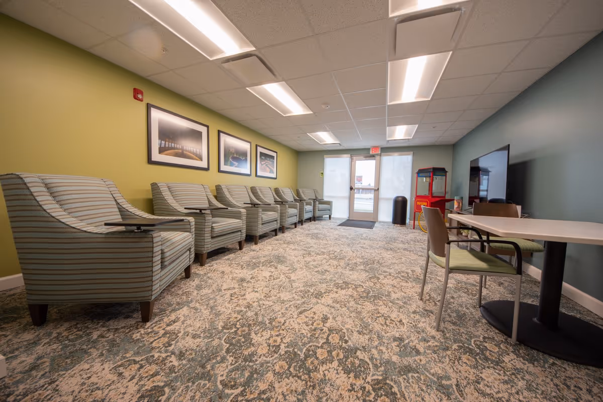 A spacious senior-living common room with a row of striped armchairs facing a TV, tables, and a popcorn machine.