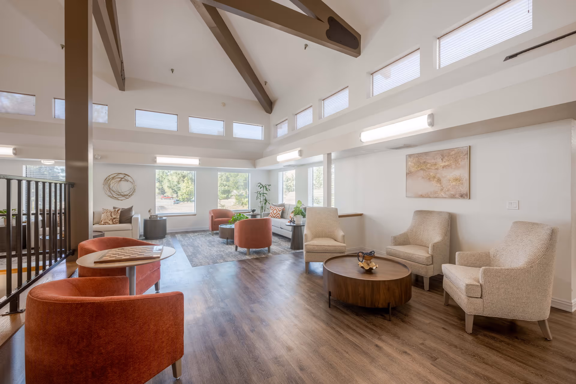 A bright and spacious living room area with high vaulted ceilings featuring exposed beams. The room is furnished with several comfortable chairs in beige and orange tones, a round wooden coffee table, and a small table with a chessboard. Large windows and clerestory windows allow natural light to fill the space. The floor is wood, and there is a decorative wall piece and a potted plant near the windows.