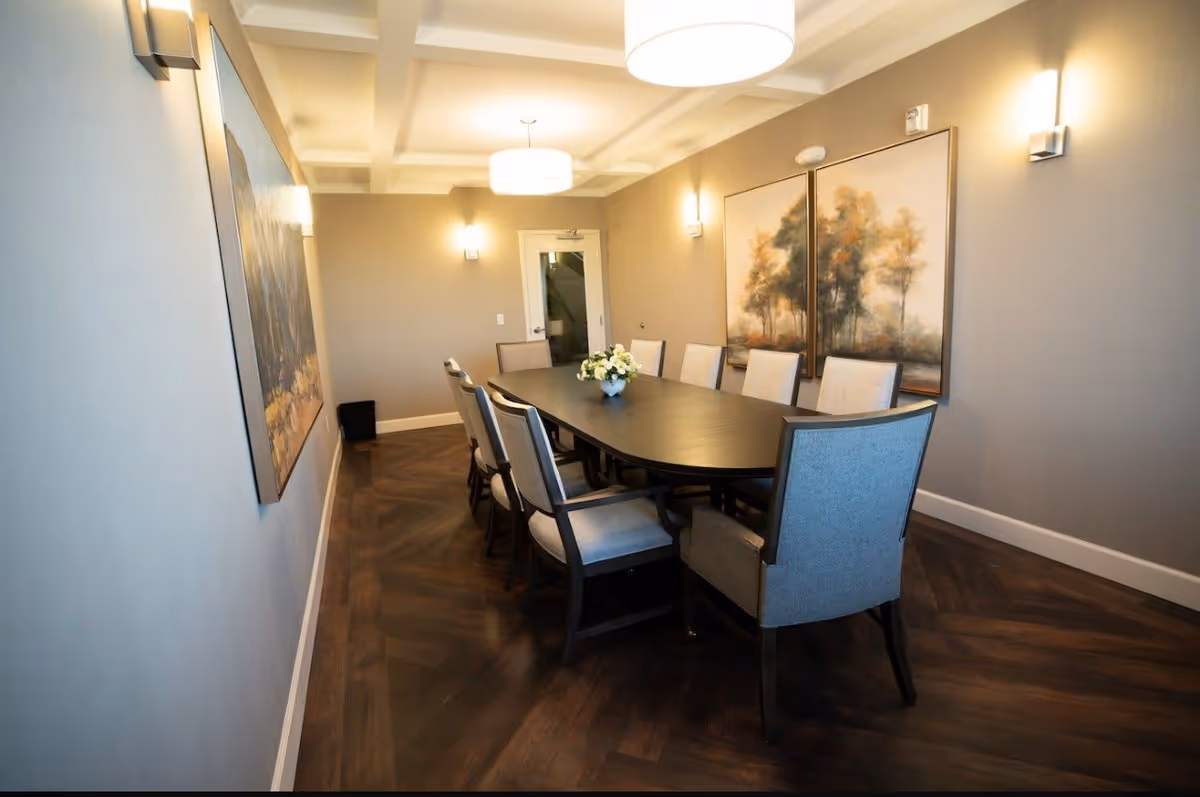 A modern dining room with a long dark wooden table surrounded by eight upholstered chairs. The room has soft beige walls, two large paintings of trees on one wall, and a coffered ceiling with two round pendant lights. The floor is dark wood with a herringbone pattern.