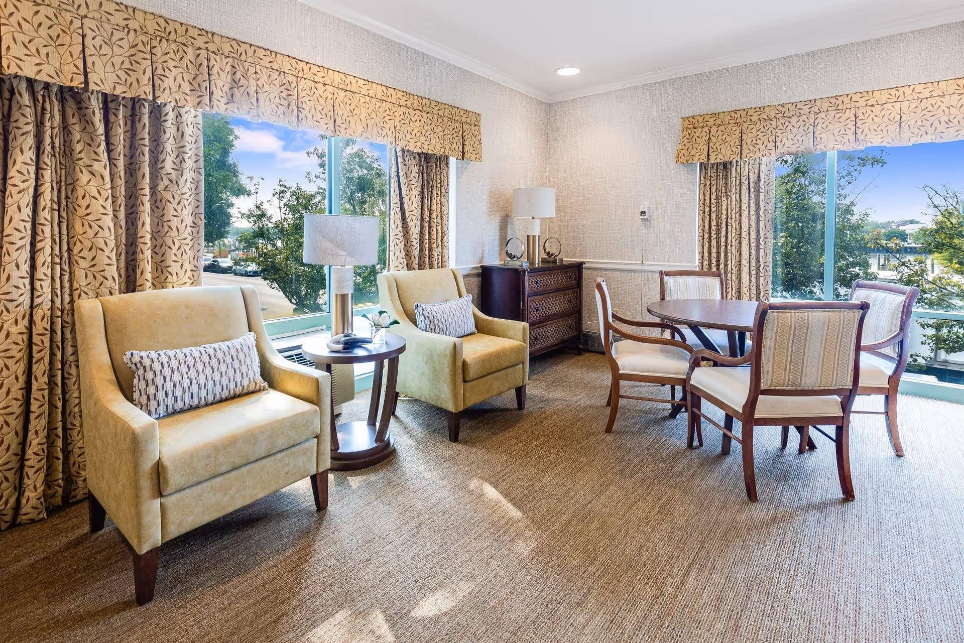 Bright common living area with two armchairs, a round dining table with four chairs, side tables and large windows overlooking trees.