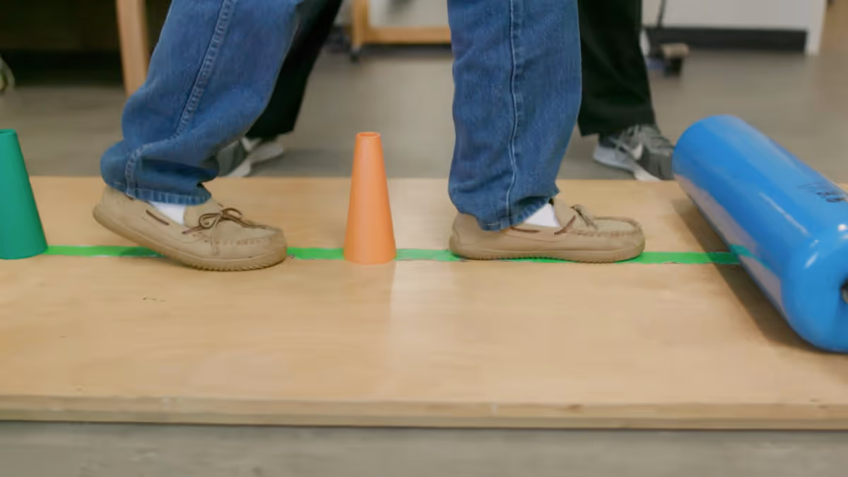 Close-up of a person's feet wearing moccasin-style shoes and jeans stepping on a wooden board with small orange and green cones and a blue cylindrical object nearby, likely part of a physical therapy or balance exercise.