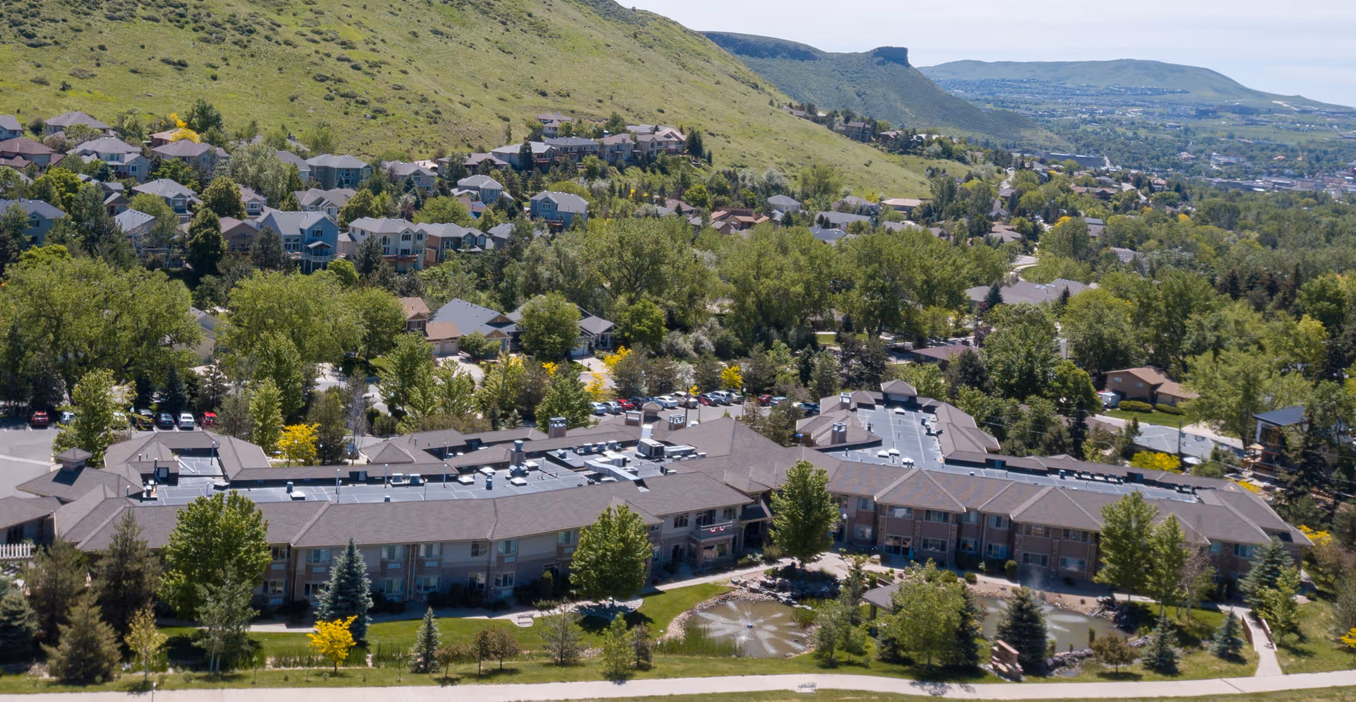 Aerial view of the Golden Pond Senior Living complex showing connected buildings, landscaped grounds with a pond, and the surrounding hillside neighborhood.