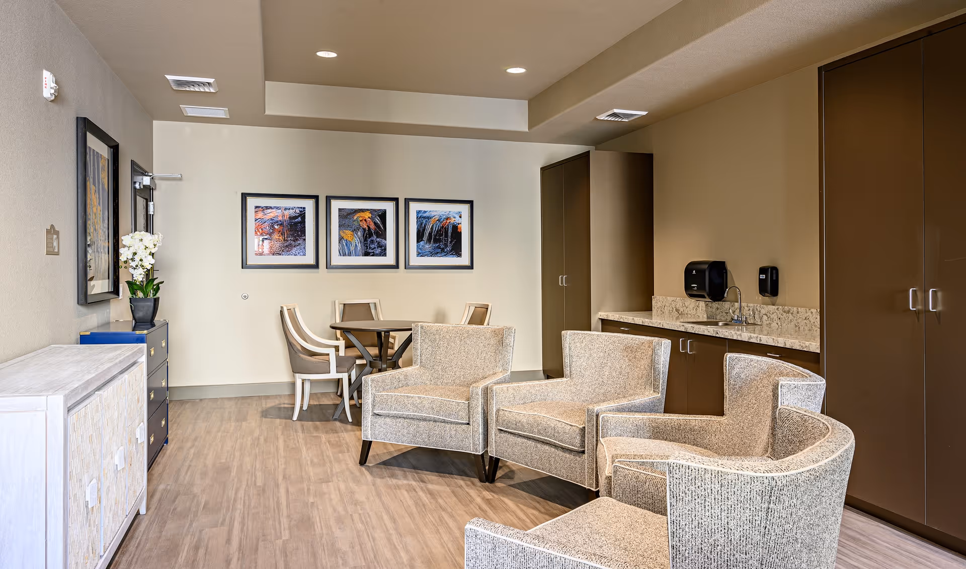 A bright communal lounge with upholstered armchairs, a small dining table, framed artwork on the wall, and a kitchenette with cabinets.