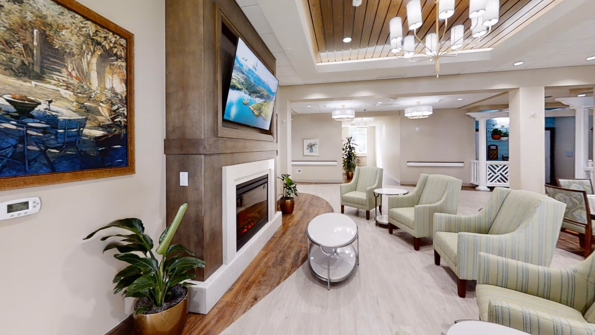 A cozy living room area in a senior living facility featuring a modern electric fireplace with a mounted TV above it. The room has light-colored walls, a wooden ceiling accent with recessed lighting, and a chandelier. There are several striped armchairs arranged around small white tables, a potted plant near the fireplace, and framed artwork on the walls.