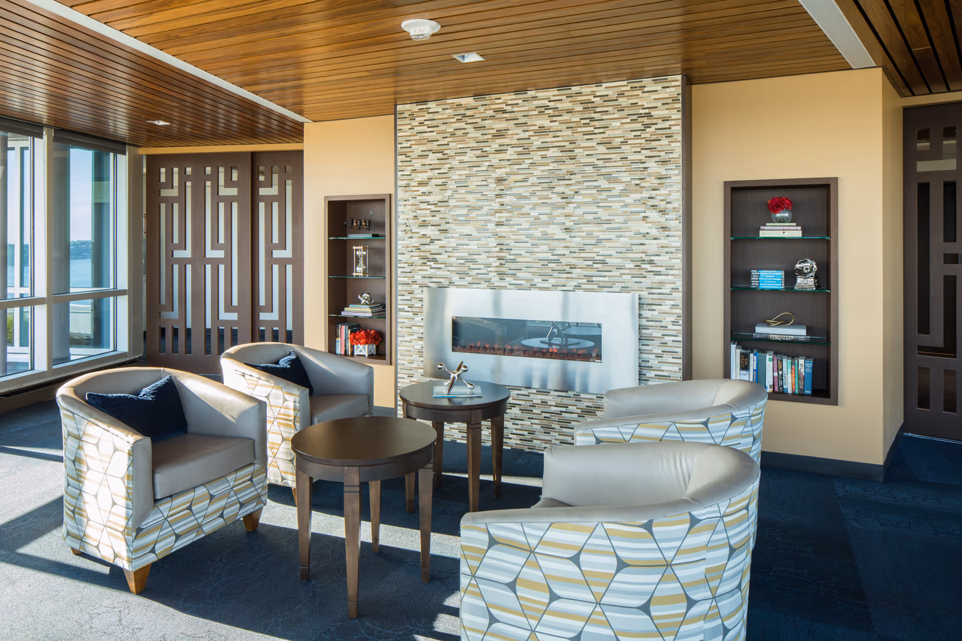 A bright communal lounge with four patterned armchairs around small tables facing a modern wall-mounted fireplace and built-in shelving.