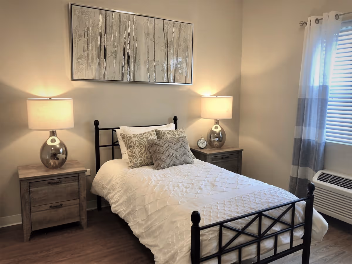Sunlit bedroom with a single bed, two bedside tables with lamps, decorative pillows, wall art, and a window with curtains.