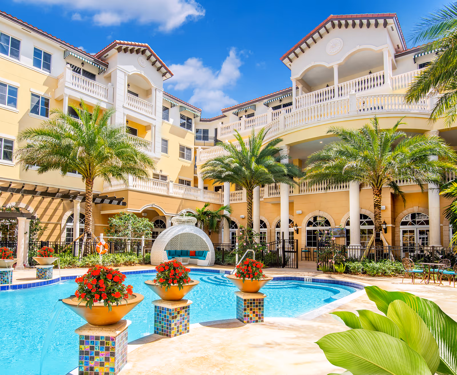 Outdoor swimming pool area at The Palace at Weston with clear blue water, decorative fountains with red flowers, palm trees, and a large multi-story building with balconies and arches in the background under a bright blue sky with some clouds.