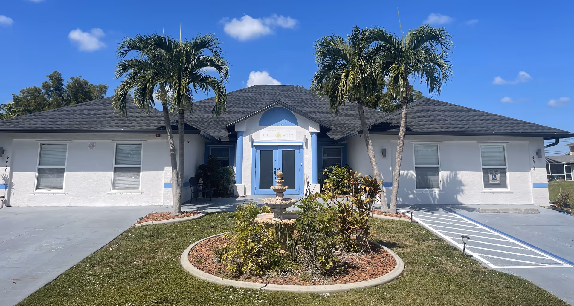 Front exterior of a single-story white assisted living building with blue double doors, two palm trees, and a small fountain in a circular landscaped bed.