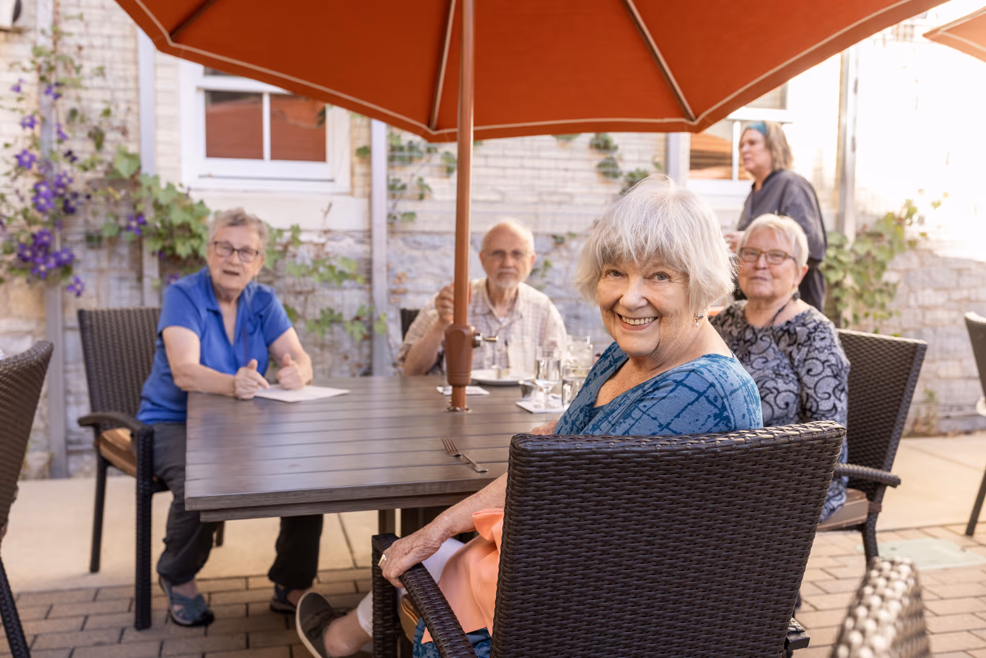 A group of elderly people sitting around a table under a large orange patio umbrella outdoors. They are smiling and appear to be enjoying a social gathering in a courtyard with brick walls and some greenery.
