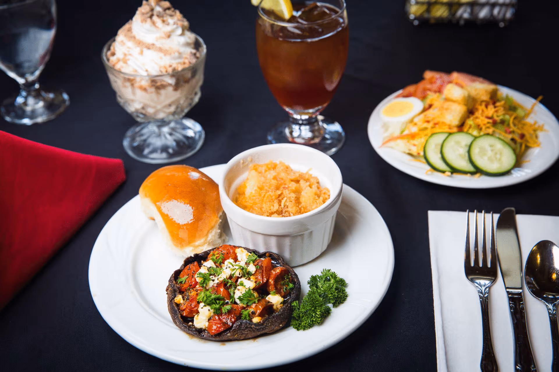 A plated meal on a white plate featuring a bread roll, a baked dish topped with herbs and cheese, and a small ramekin of a baked side dish. Next to the plate is a fork, knife, and spoon on a white napkin. In the background, there is a glass of iced tea with a lemon wedge, a dessert cup with whipped cream and nuts, a water glass, and a plate with salad including cucumber slices, a boiled egg, and croutons, all set on a black tablecloth with a red napkin.