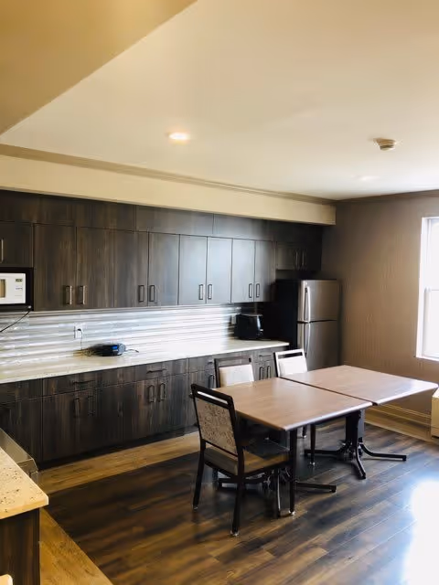 A kitchen area with dark wood cabinets, a countertop with a microwave, coffee maker, and a refrigerator. There is a wooden table with four chairs in the center of the room. The room has wooden flooring and a window letting in natural light.