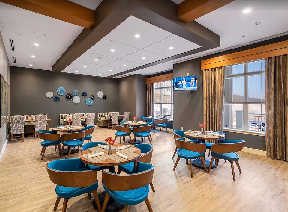 A bright and modern dining room with round wooden tables surrounded by blue cushioned chairs. The room features large windows with beige curtains allowing natural light to fill the space. On one wall, there are decorative plates in shades of blue and white. A flat-screen TV is mounted on a gray wall near the windows. The floor is light wood, and the ceiling has recessed lighting with a geometric design.
