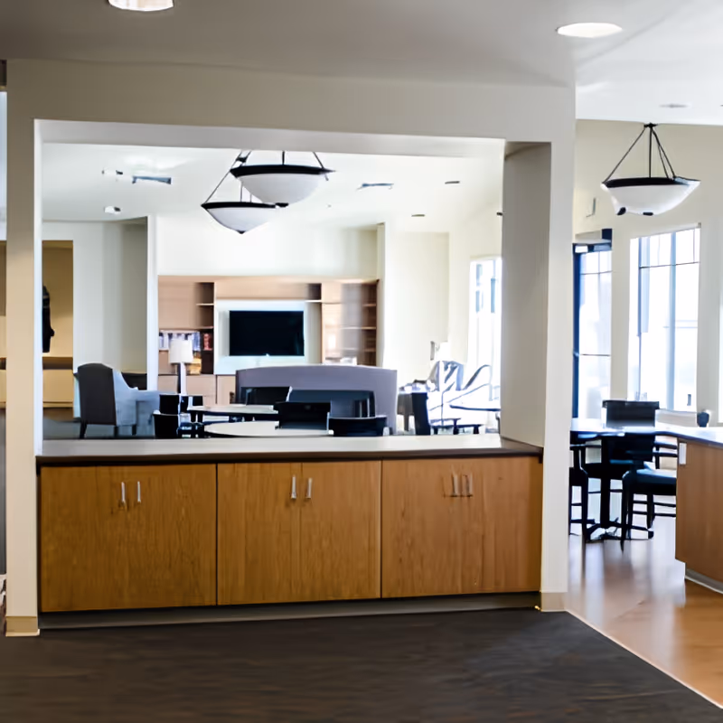Interior view of a senior living facility common area with wooden cabinets in the foreground, modern light fixtures hanging from the ceiling, a seating area with chairs and a TV in the background, and large windows letting in natural light.