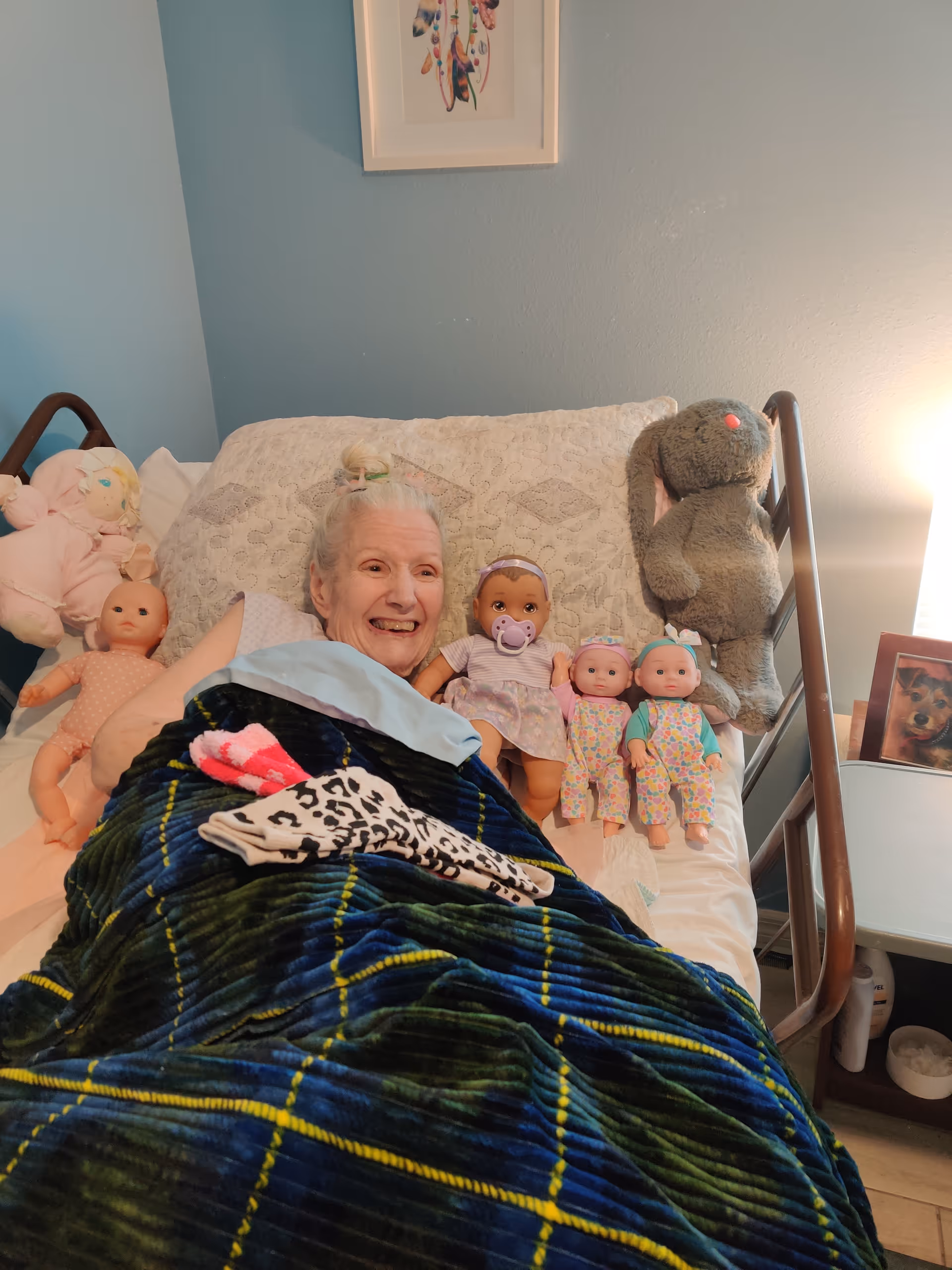 An elderly woman lying in a bed, smiling and surrounded by dolls and stuffed animals in a cozy bedroom.