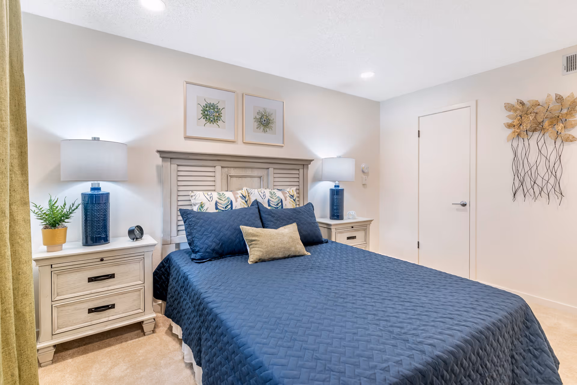 A neatly made bed with a blue quilt and decorative pillows in a bright bedroom. The bed has a wooden headboard and is flanked by two matching nightstands, each with a blue lamp and decorative items. There are two framed botanical prints on the wall above the headboard, a potted plant on the left nightstand, and a metal wall decoration on the right wall next to a closed door.