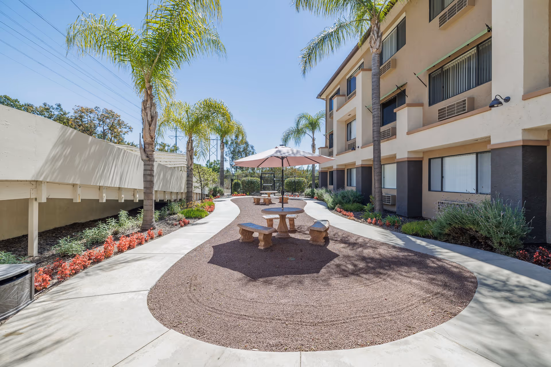 Outdoor courtyard area at Rancho Penasquitos Senior Living with palm trees, a gravel seating area featuring round stone tables and benches under umbrellas, surrounded by a concrete walkway and landscaping with red flowers and green shrubs, adjacent to a multi-story building.