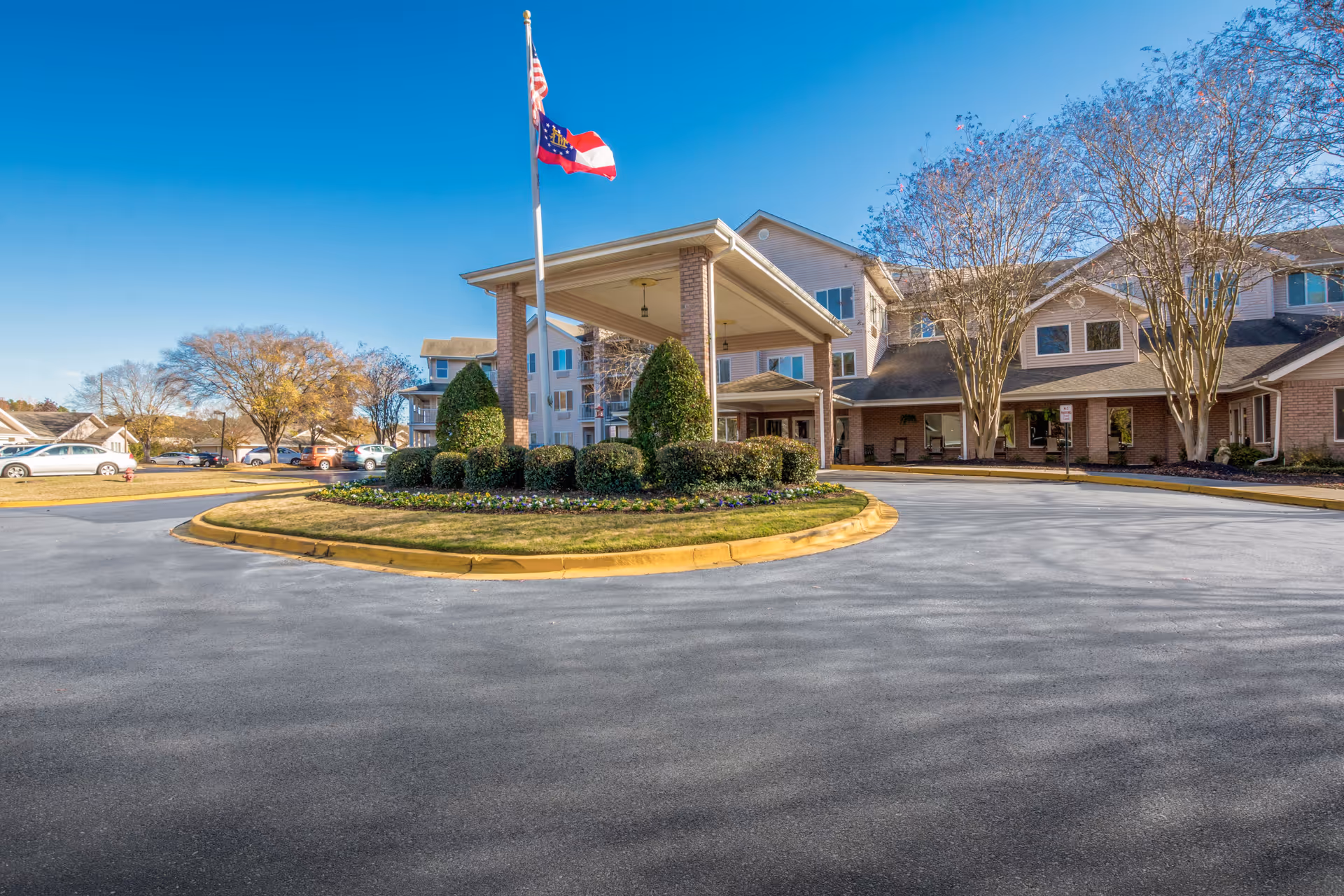 Front exterior view of Washington Commons by Barclay House, showing a circular driveway with a landscaped island in the center featuring bushes and a flagpole with the American and Georgia state flags. The building is a multi-story structure with a covered entrance and several windows, surrounded by trees and parked cars.